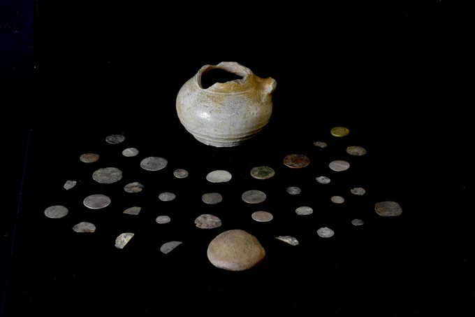 The vessel containing the 36 coins appears to be a European stoneware vessel, perhaps previously a jug, which was repurposed to store the coins. The stone lid appears to be a reused polishing stone for leatherworking. 