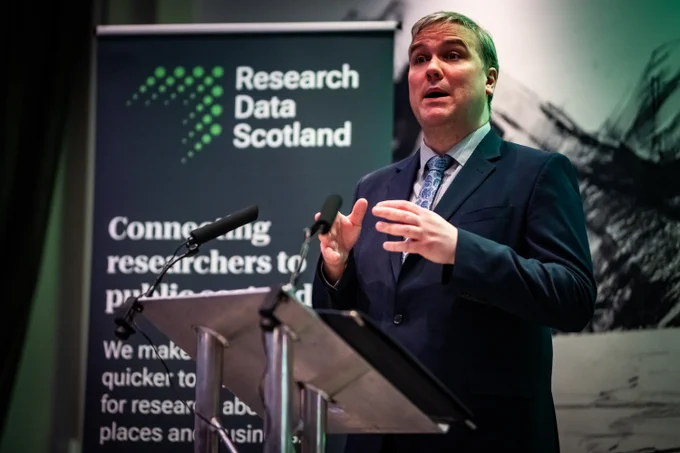 A man giving a speech at a metal lectern. A banner in the background displays the Research Data Scotland logo.