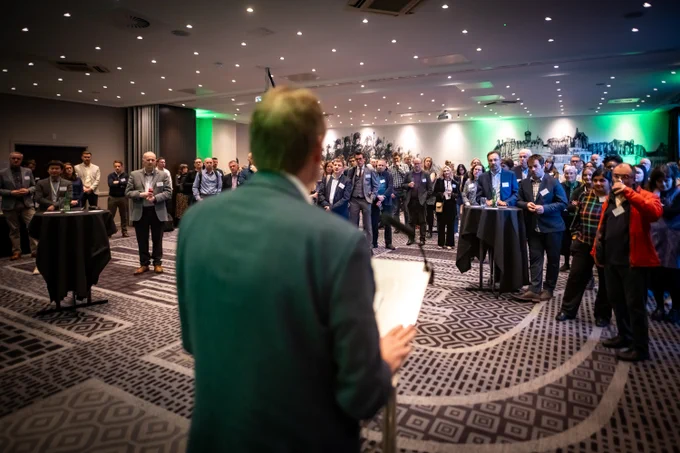 A room of people stand listening to a speech. In the foreground, a man faces away from the camera, speaking to the audience.