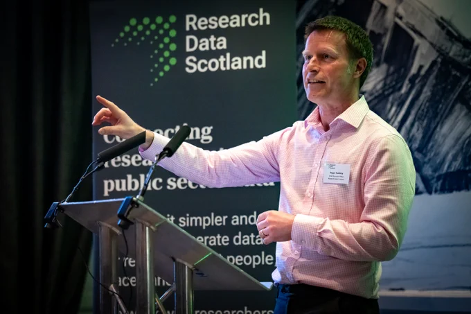 A man giving a speech at a metal lectern. A banner in the background displays the Research Data Scotland logo.
