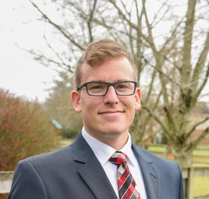 Man in blue suit with red, gray and black tie and black rectangular glasses smiles at the camera. You can see trees behind him. 
