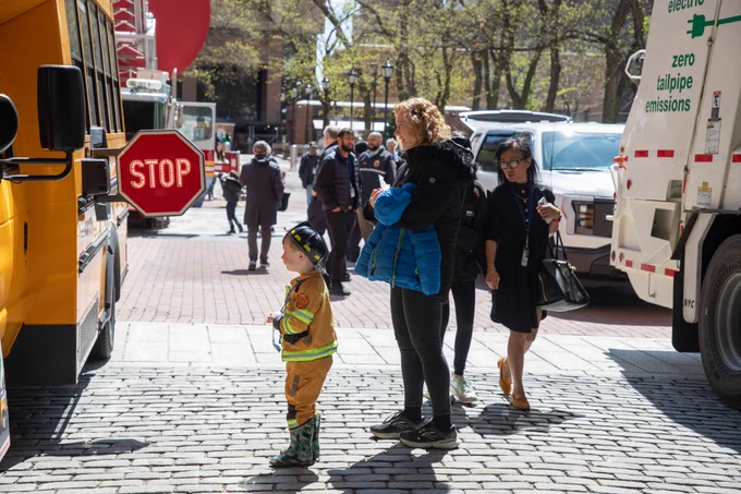 Young child wearing a firefighter uniform stands with his parent observing an electric school bus. 