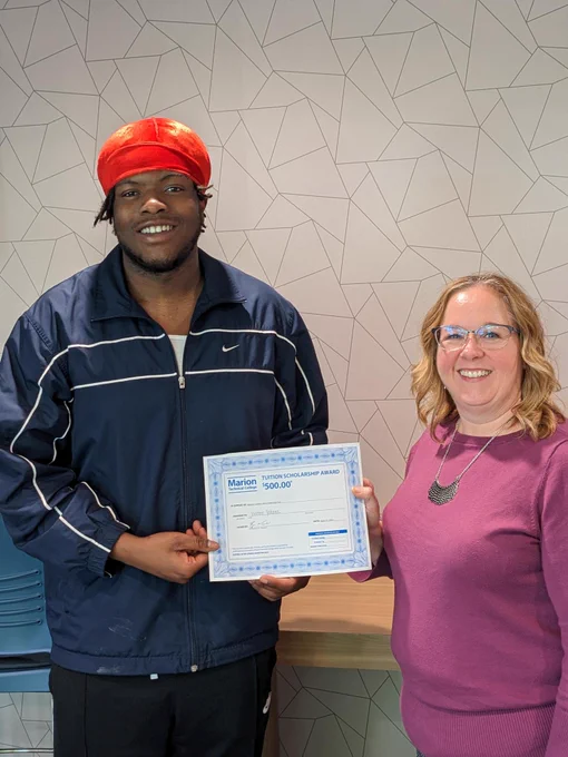 A student holds a $500 scholarship certificate and smiles while standing next to a staff member. 