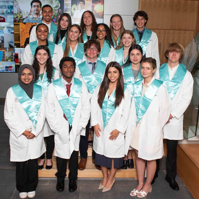 Senior high school students wearing white coats with teal collars pose in front of a science media collage.