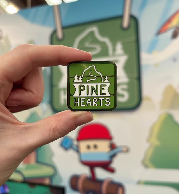 A hand holds a pin badge of the Pine Hearts logo up in front of the Pine Hearts booth artwork at WASD.