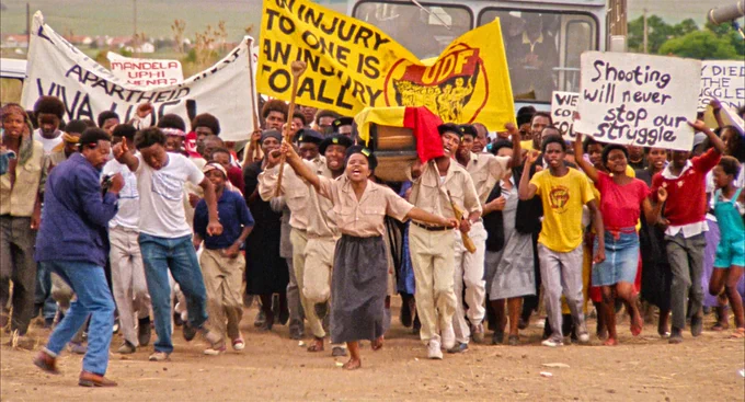 Still from the film Mapantsula. A big group of black people protesting with banners, one saying an injury to one is an injury to all. Another banner says shooting will never stop our struggle. A bus is in the background. A woman is leading the group defiantly 