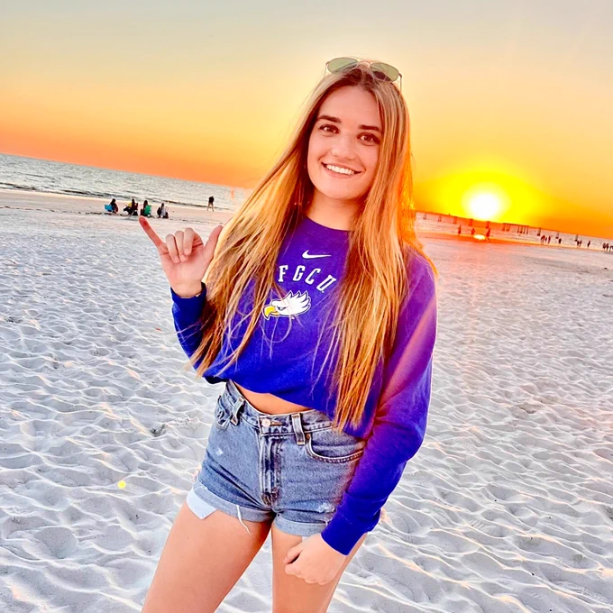 a student stands on a beach with an FGCU hoodie and a wings up
