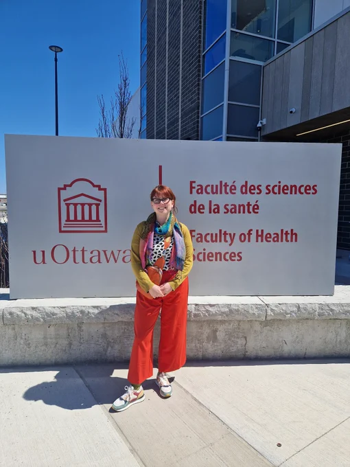 Tai standing in front of university ottawa sign wearing bright red trousers and smiling