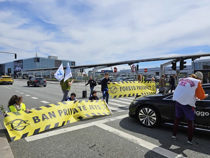 Activists blocking the road in front of Copenhagen/Kastrup airport holding banners written "ban private jets". A car is stopped in front of the banners. 