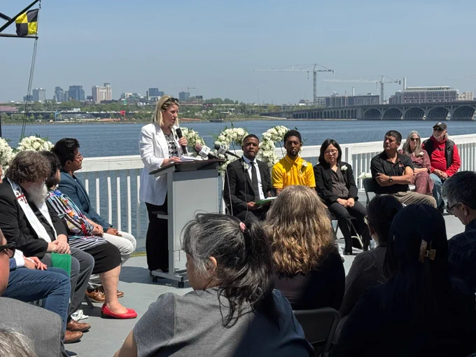 Photo of AFL-CIO President Shuler speaking at a podium with a group of other dignitaries seated next to her and an audience in front of her. She is speaking outside from a deck with the Baltimore skyline behind her. 