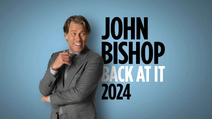 Man in suit with arms crossed, smiling looking off camera. Text reads JOHN BISHOP BACK AT IT 2024