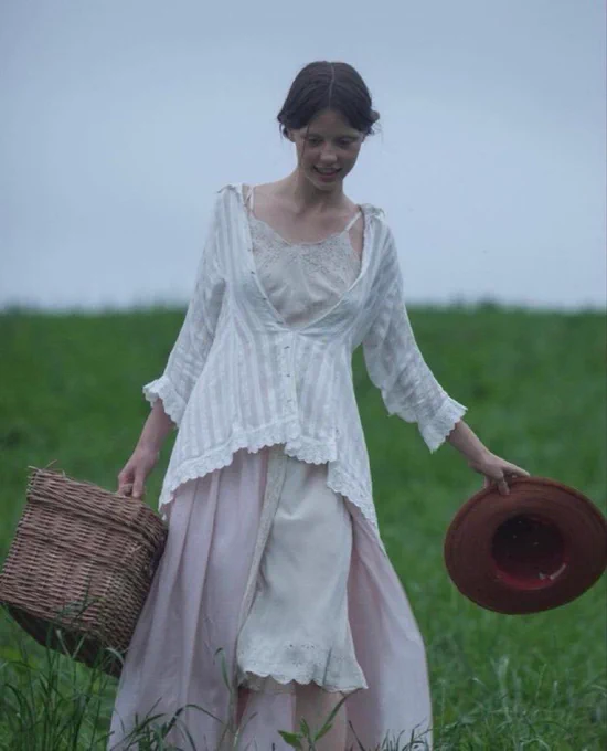 Mia Goth vestida de época anda por una pradera mientras lleva en una mano una cesta para el pícnic y en la otra un sombrero de paja.