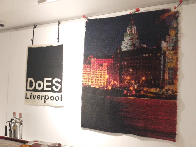 Photo showing two rag rugs hanging on a studio wall. One of the rugs incorporates the DoES Liverpool logo in black & white. The other larger rug depicts the Liverpool waterfront at night. 