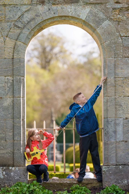 A photo of two children in a historic stone archway. One child is sitting down holding a tape, the other is reaching up with the end of the tape to take a measurement.