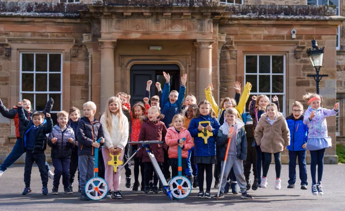 A photo of a class of children in front of Newbattle Abbey College. Children in front holding surveying equipment, children behind jumping up.