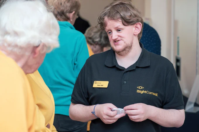 A man wearing an iSightCornwall branded polo shirt is talking to a person wearing a yellow jumper