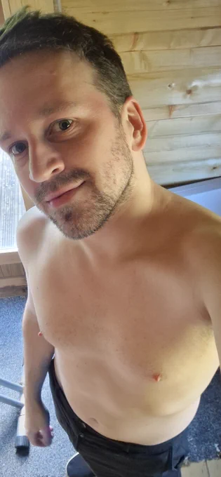 A gym selfie of a topless white cis Scottish guy with brown hair (short back and sides), brown eyes, and a bit of a beard. Wearing black trousers in a wooden shed.
