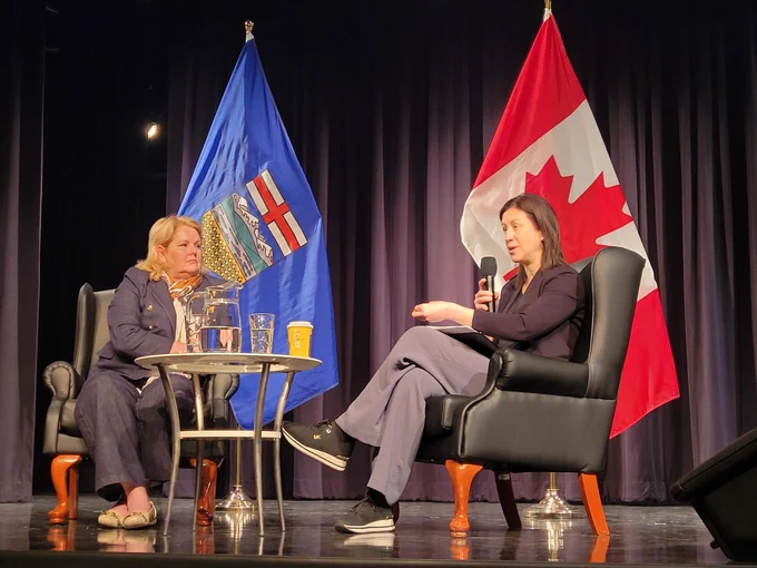 Deputy Clerk Fox speaks on stage seated next to Michele Evans, ADM PrairiesCan. Canada and Alberta flags in the background.