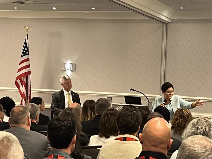 The U.S. Fire Administrator speaks into a microphone in front of an audience with one arm extended, as CFSI Executive Director, Bill Webb, asks questions. A U.S. flag stands to the left of Mr. Webb. 