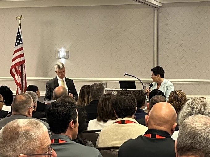 The U.S. Fire Administrator speaks into a microphone in front of an audience as CFSI Executive Director, Bill Webb, asks questions. A U.S. flag stands to the left of Mr. Webb. 