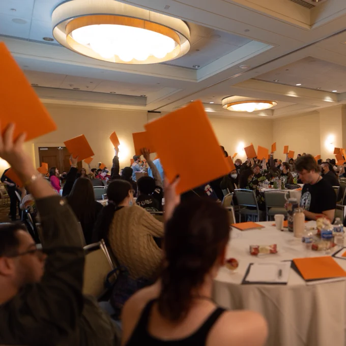A large group of Starbucks Workers United members sit in a ballroom and raise orange folders in the air