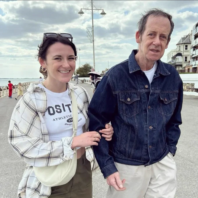 A woman (left) and older man (right smiling at the camera. The woman is holding onto the man's arm. They are stood outside.