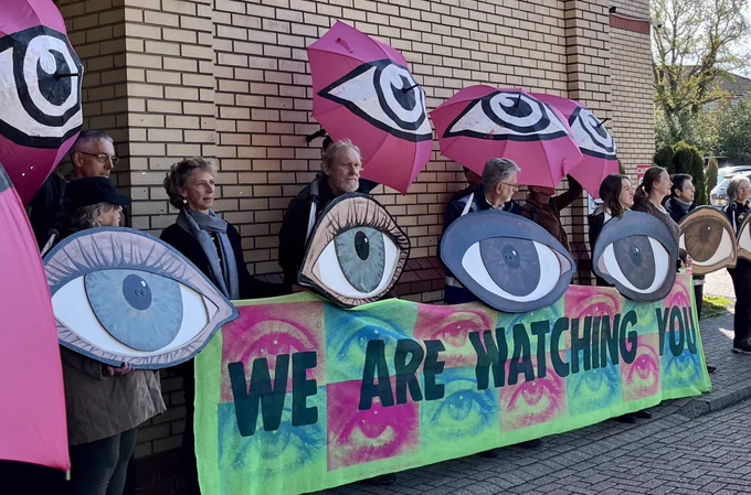 Group of XR campaigners outside the hotel hosting the Gatwick expansion examination hearing, holding up giant eye placards and a banner saying ‘We are watching you’.