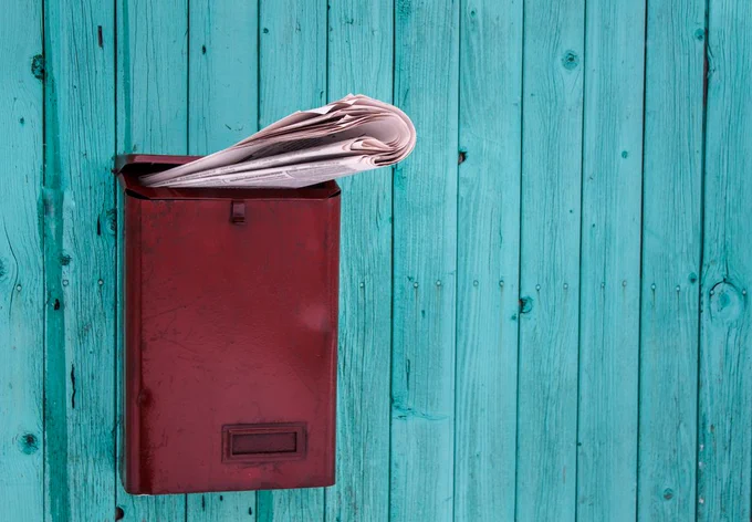 A rolled-up newspaper sticking out of a red letterbox on a turquoise wooden fence. Image by AVAKAphoto via Shutterstock.