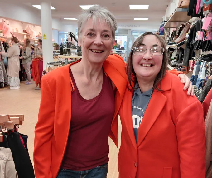 Kath and Annaliese in matching orange jackets in a charity shop with their arms around each other's shoulders