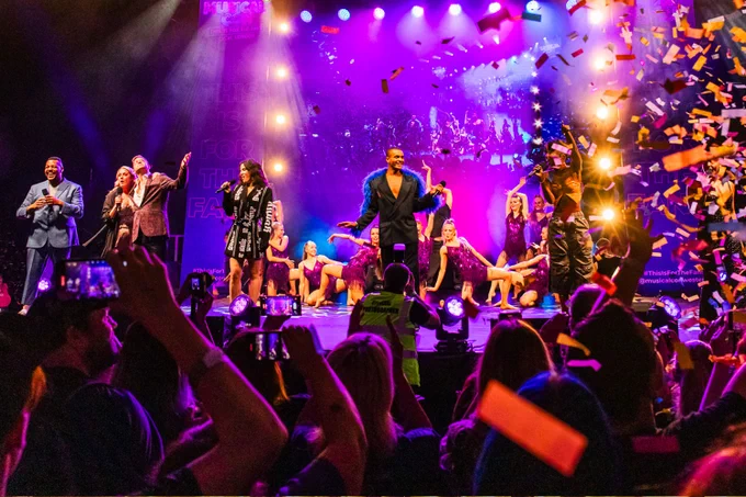 A photo showing a group of performers on a raised dias surrounded by fans cheering