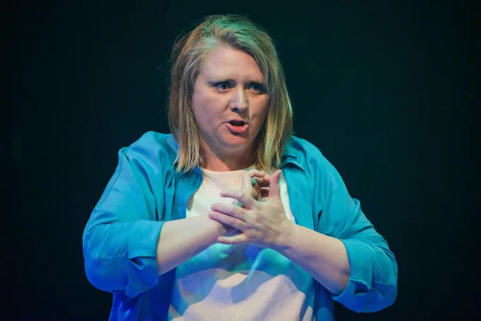 Production shot of Trine Garrett captured mid-speech, animated and rubbing her hand. She has blonde hair and is wearing a bright blue shirt with a white t-shirt underneath.