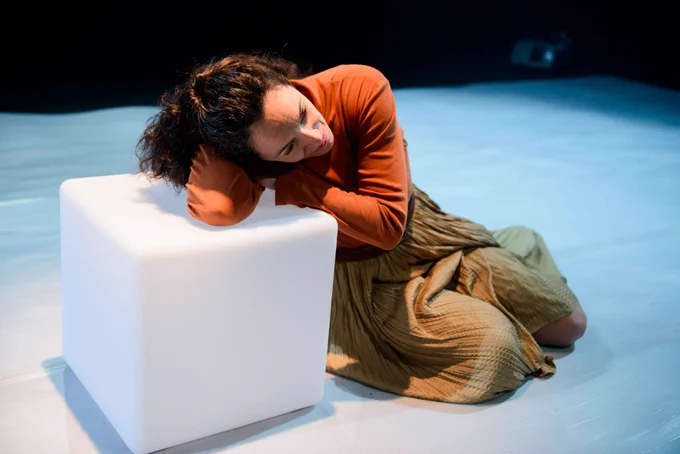 Production shot of Camila França sitting on the floor, leaning on a cube bathed in light. She has long brown hair and is wearing a burnt orange top and a green skirt.