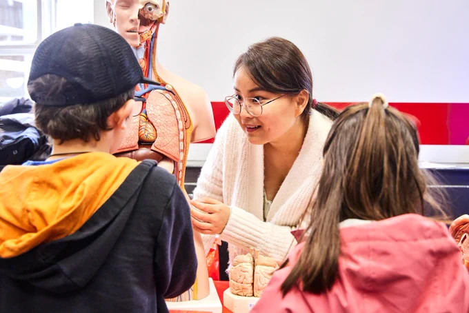 Two children talking to an academic, who is pointing to a human body dummy 