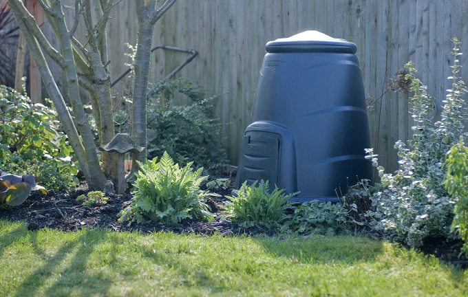 A plastic compost bin inthe sunshine in a flower bed by a lawn.