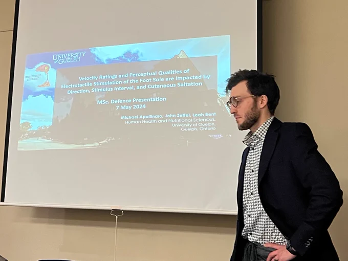 A photo of Michael at his defence, in front of a projected title screen with the same title of his thesis that is listed in this posting.