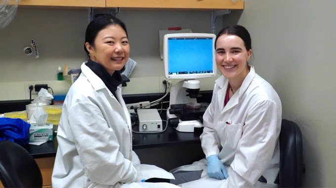 Graduate students Clara Xia and Stephanie Besoiu sit in front of a microscope on a lab bench in the Jefferies lab.