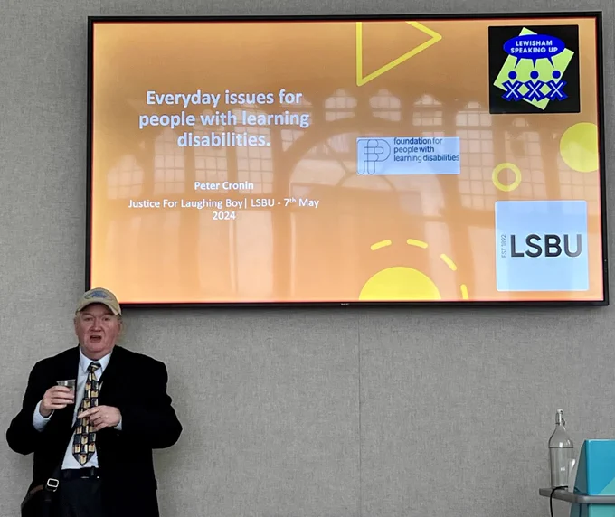 Photo of Peter presenting, baseball cap, suit and tie standing beneath a large screen.