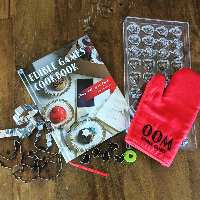 An Edible Games Cookbook surrounded by an oven mitt, chocolate mould, vegetable cutters, and cookie cutters.