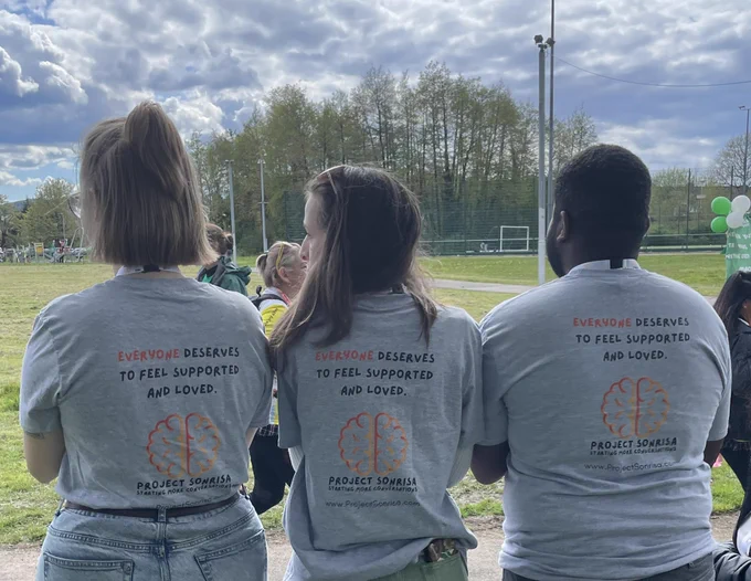 Three young people with their backs to the camera. Each is wearing a grey t-shirt with text on the back reading ‘Everyone deserves to feel supported and loved’.