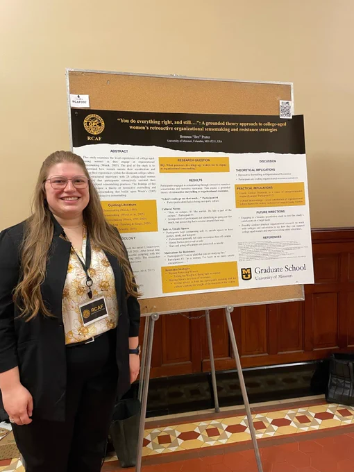 Bre Prater is standing in front of her research poster. The poster is white with black and gold accents. Bre is wearing a white and gold blouse and a black blazer. Her hair is down and she wears glasses.