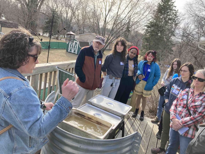 A group of people stand outdoors on the deck of a house. A woman with a denim jacket and sunglasses shows them the trays of tree sap that she has boiling on a propane stove, protected from the wind by a curved piece of corrugated metal. This will eventually be boiled down into maple syrup.