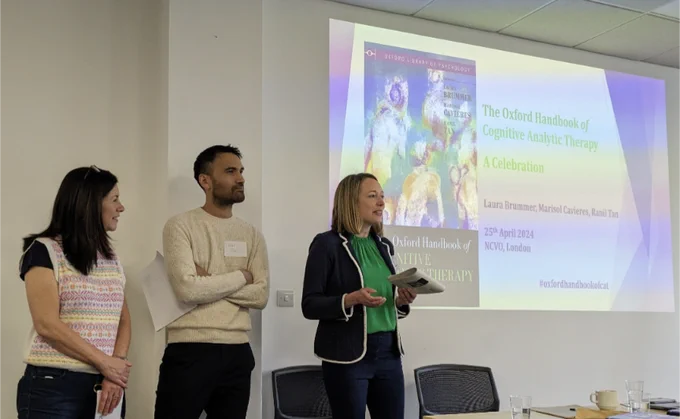 Marisol Cavieres and Ranil Tan stand next to Laura Brummer as she talks in front of a slide introducing the Oxford Handbook of Cognitive Analytic Therapy - A Celebration