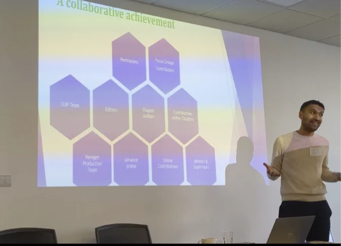 Ranil Tan talking, arms open, in front of a slide showing ten purple hexagons with text describing the many collaborations that helped bring the Oxford Handbook of CAT to fruition.