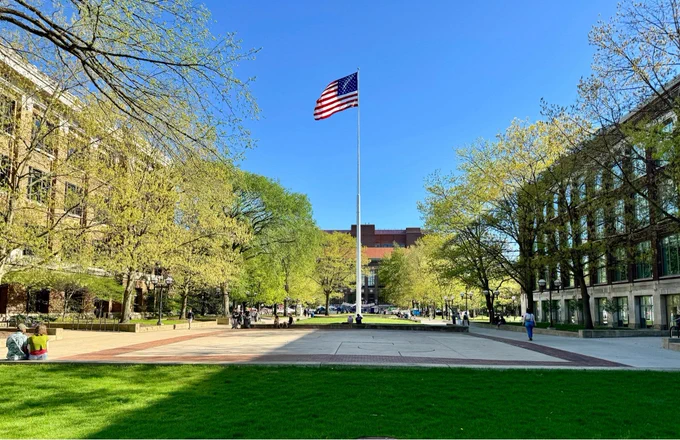 Outdoor photo of the University of Michigan diag. There are some people calming hanging around. The encampment is in the background. An American flag occupies the center of the image.