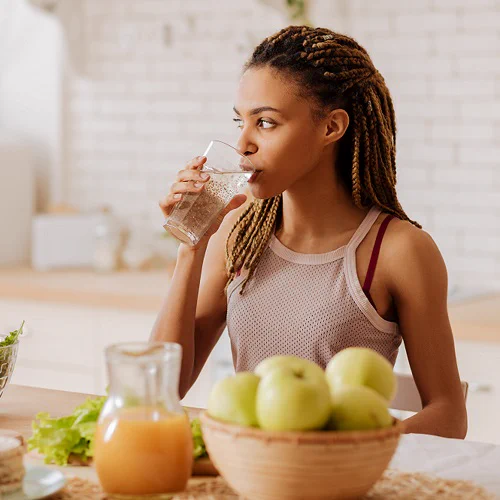 Woman drinking water with a bowl of apples, salad and fruit juice in the foreground