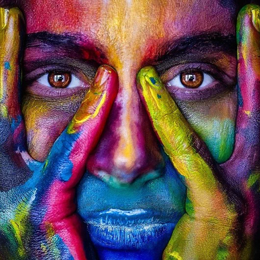 A close up of a person with their hands pressed against their face. Their skin is painted in blue, purple and yellow.