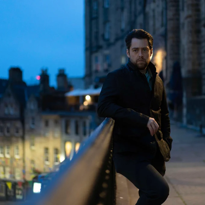 Richard Rankin as John Rebus, leaning against the railing of a balcony and looking to camera. 