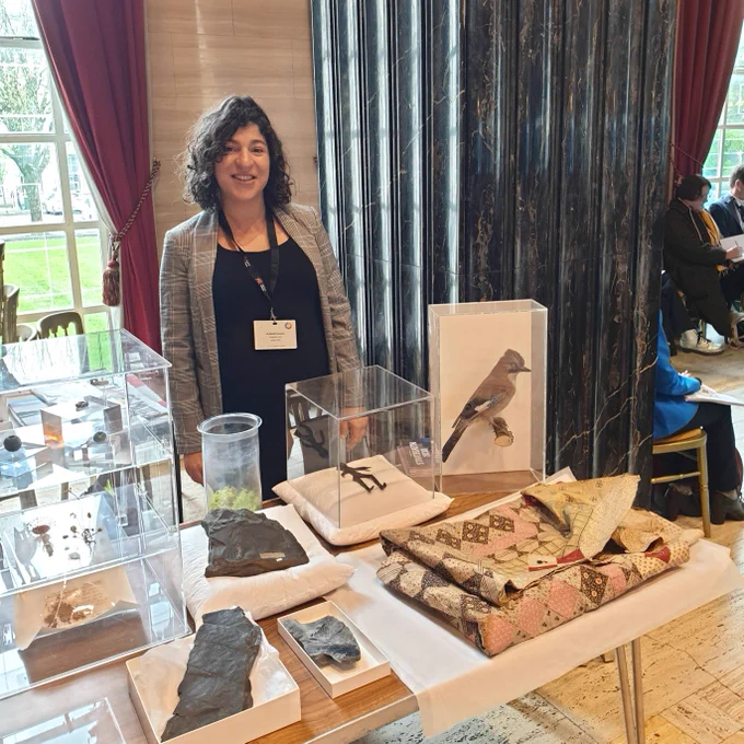 Photo of Dr Sarah Younan standing in front of a table with a number of specimens from Amgueddfa Cymru's collections. The specimens include a bird, a blanket and fossils of all kinds.