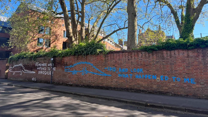 Image of the brick wall on Blackhall Road behind Keble, with graffiti of what looks like two dinosaurs. Text reads 'Remember what happened to the dinosaur!' 'I did, and look what happened to me'.