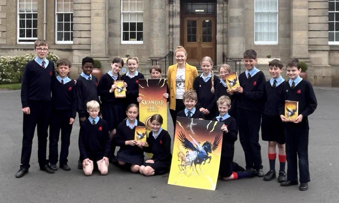 Children standing outside a school with AF Steadman in a yellow jacket with posters of unicorns from yellow chaos trials cover and holding copies of the book